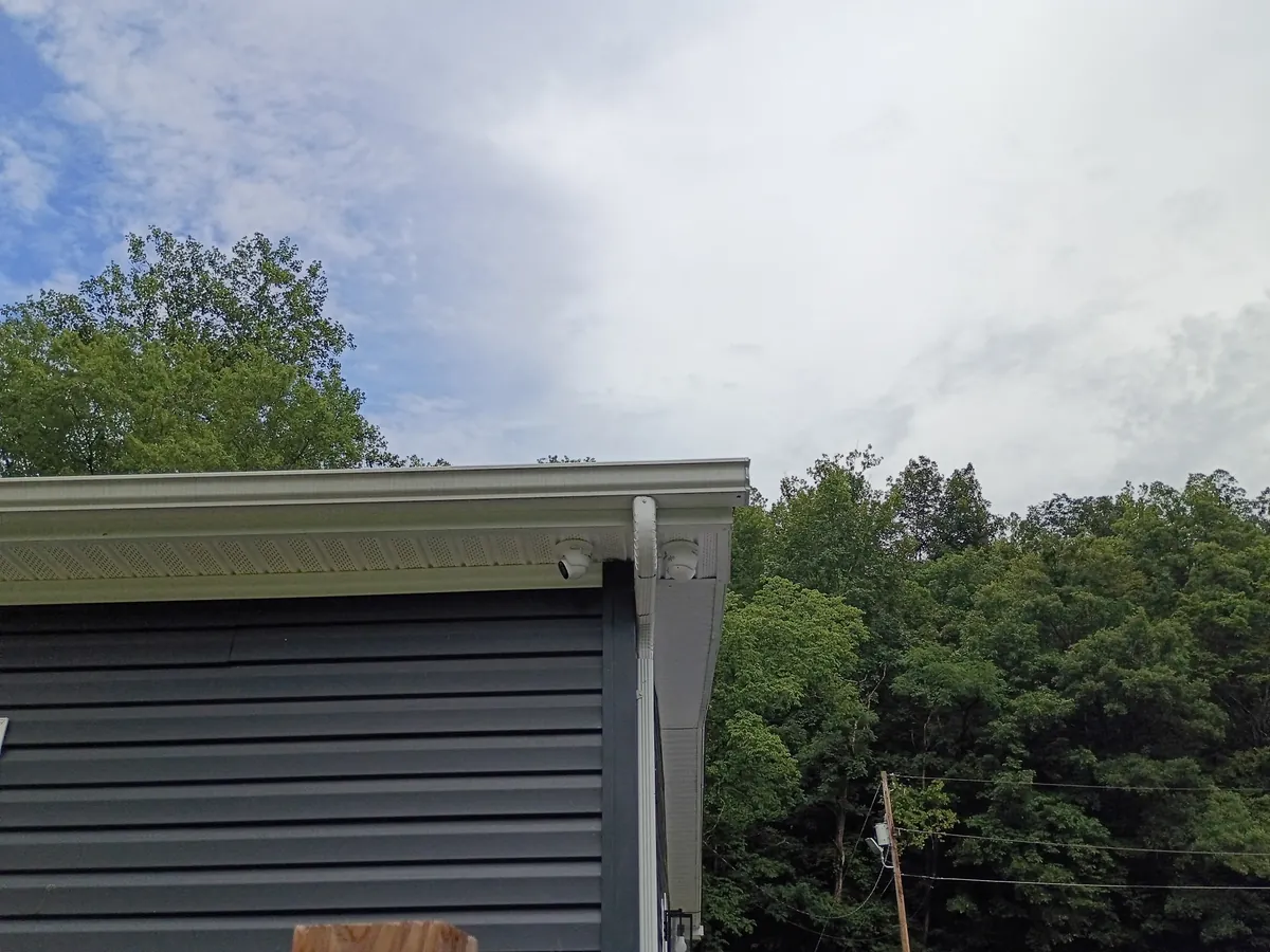 Two security cameras mounted at the corner soffit of a home overlooking a wooded yard