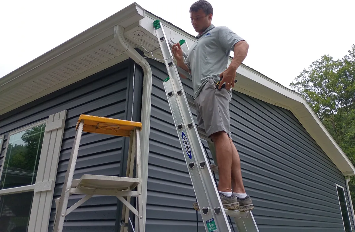An ODA installer on a ladder wiring a security camera at the gable of a home